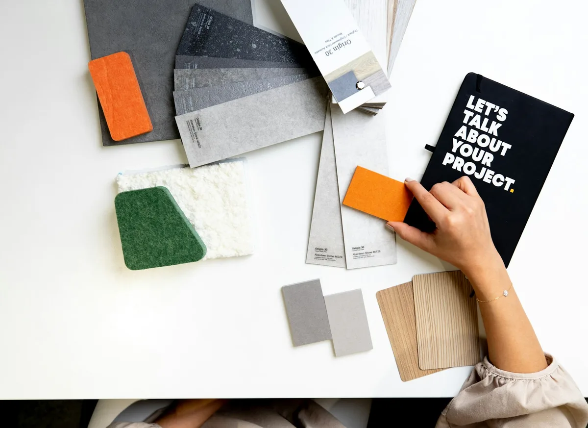 Hand holding a material swatch over a table of fabric, stone and wood samples beside a notebook